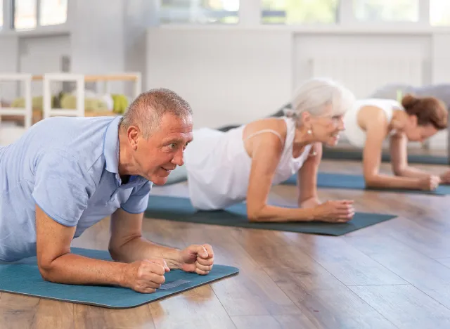 Group of seniors including an older man holding a forearm plank on yoga mats in a bright studio