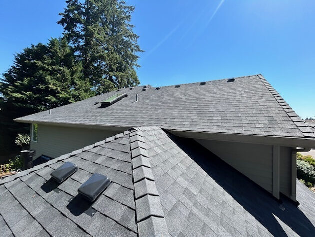 Completed architectural shingle roof with ridge caps and roof vents under a clear blue sky