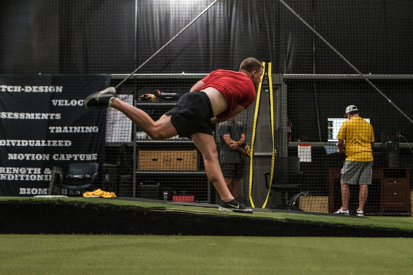 A young pitcher throwing off a mound in a training facility while coaches monitor his mechanics and workload