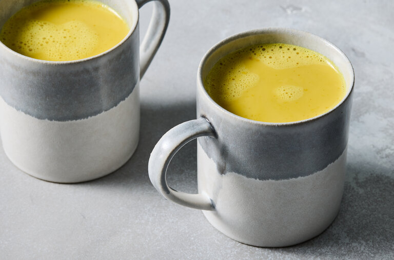 Two handmade ceramic mugs of frothy golden turmeric milk latte on a soft gray surface