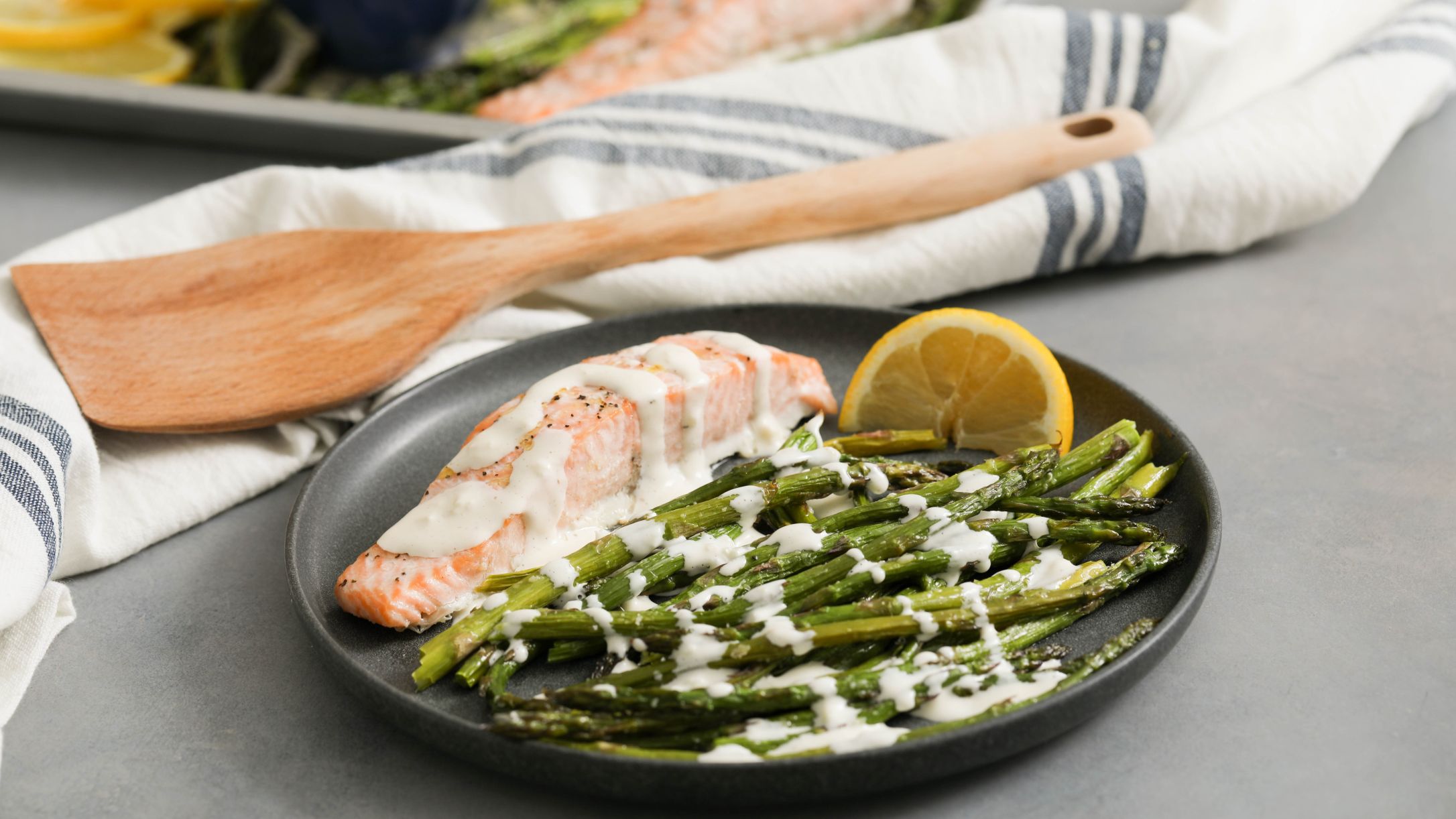 Cooked salmon fillet drizzled with creamy sauce, served with roasted asparagus and a lemon wedge on a dark plate