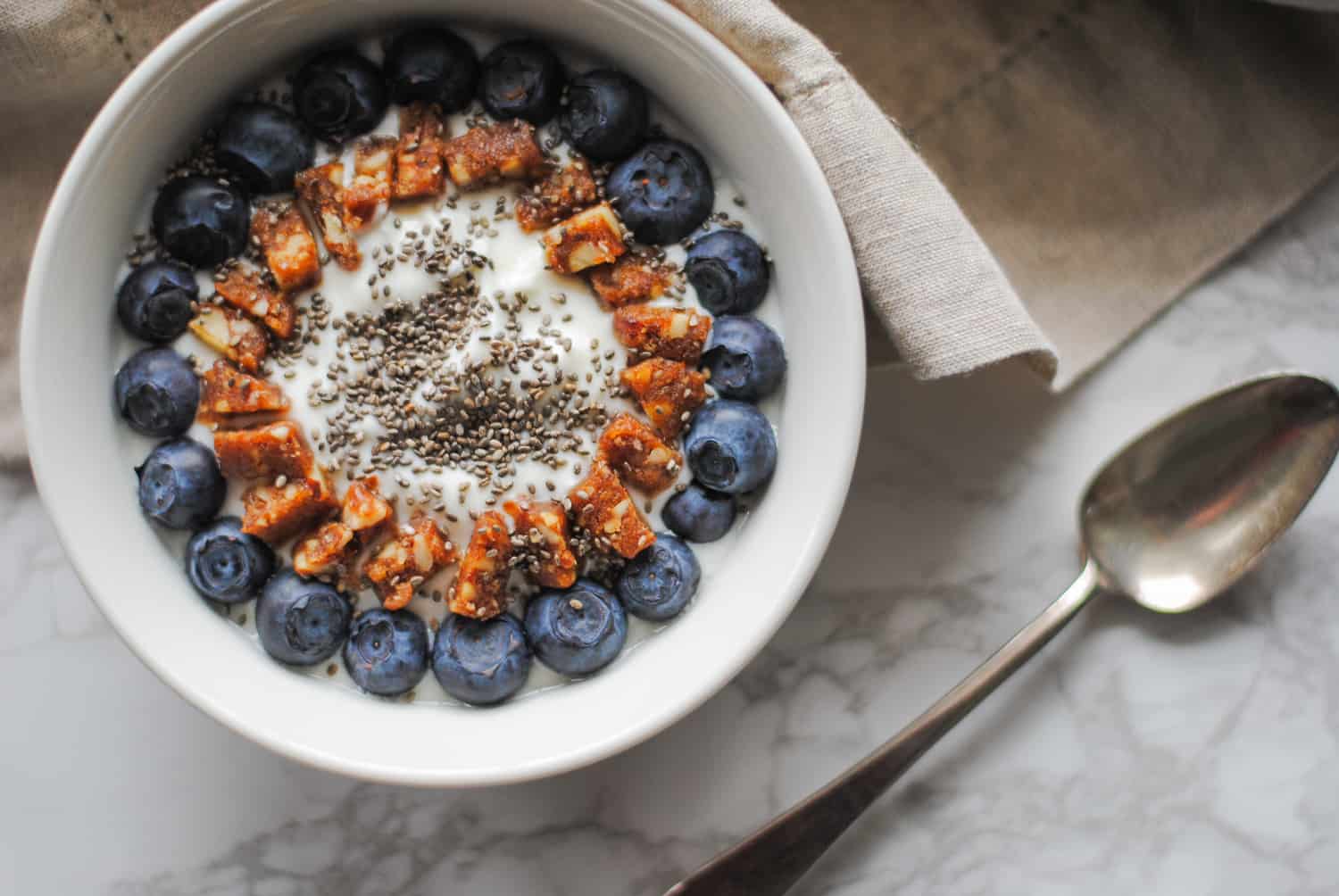 White bowl of creamy Greek yogurt topped with a ring of fresh blueberries, chia seeds and chopped date-nut bars on marble