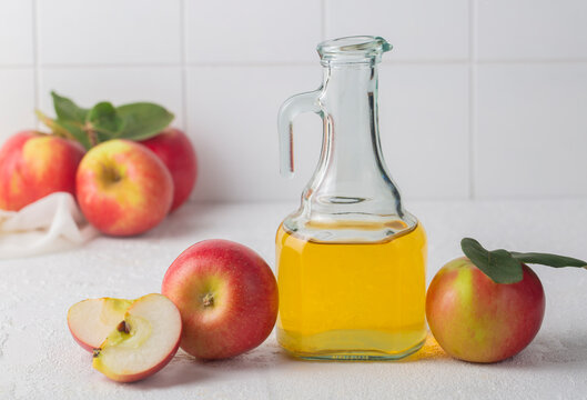 Glass cruet of golden apple cider vinegar beside fresh red apples and a cut apple half on a bright white kitchen surface
