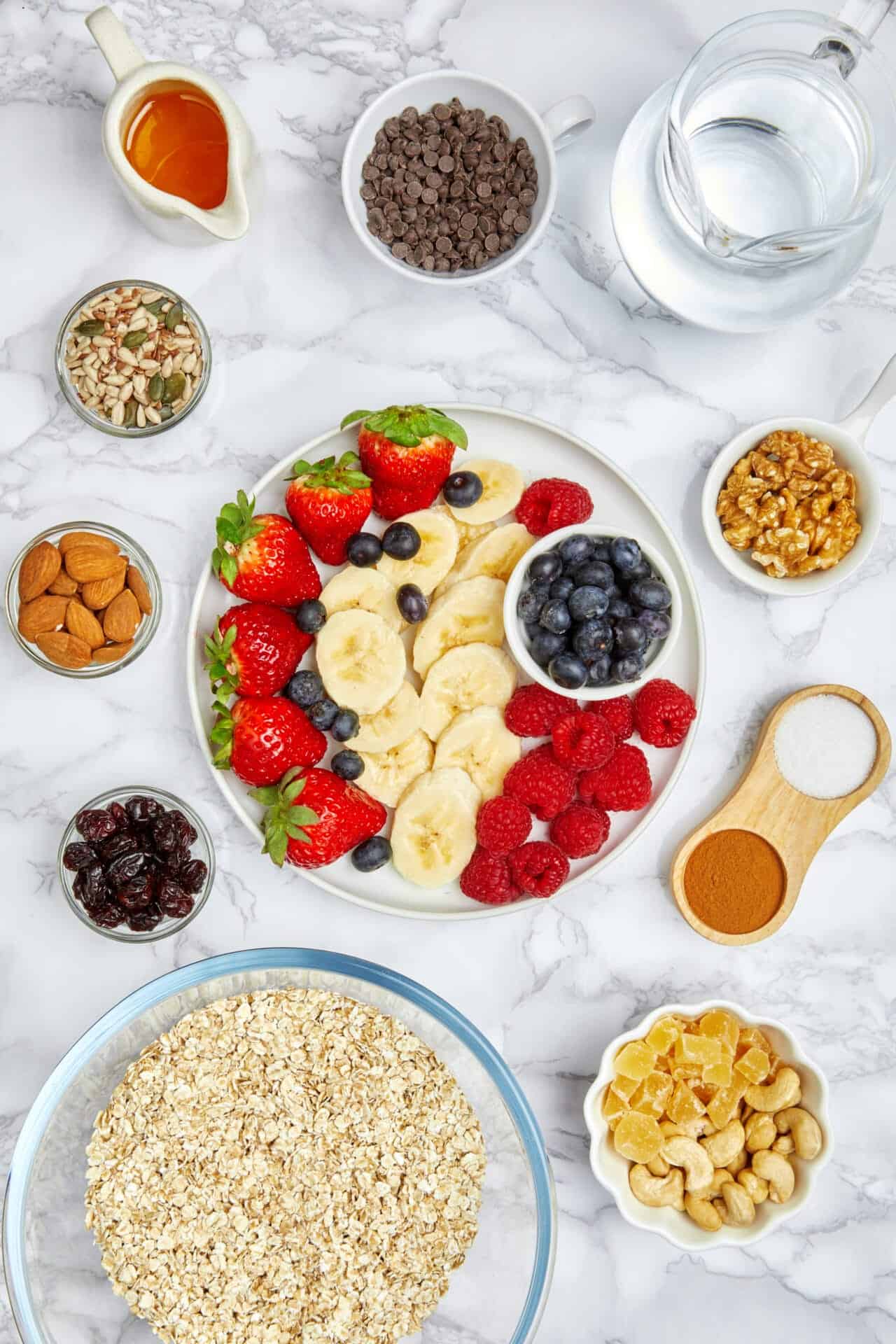 Overhead flat-lay of diabetes superfood breakfast toppings: rolled oats, strawberries, blueberries, raspberries, bananas, almonds, walnuts, pumpkin seeds, cinnamon and honey on white marble