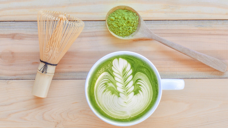 Top view of a frothy matcha green tea latte with leaf latte art, a bamboo whisk and a wooden spoon of matcha powder on light wood