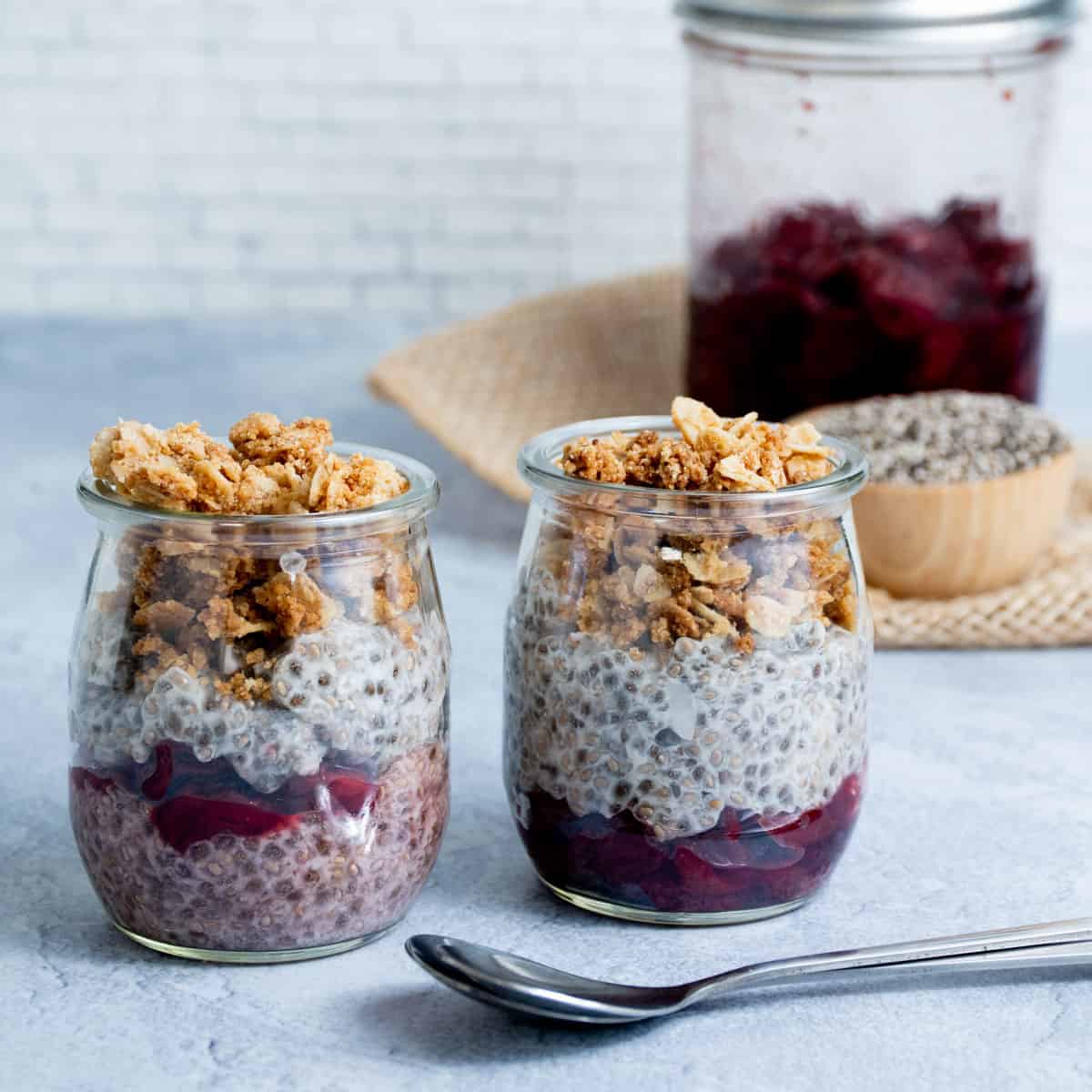 Two glass jars of layered chia seed pudding topped with crunchy granola, with a wooden bowl of dry chia seeds in the background