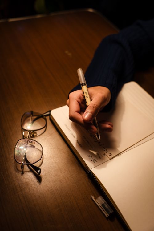 A hand writing in a journal with a pen beside reading glasses
