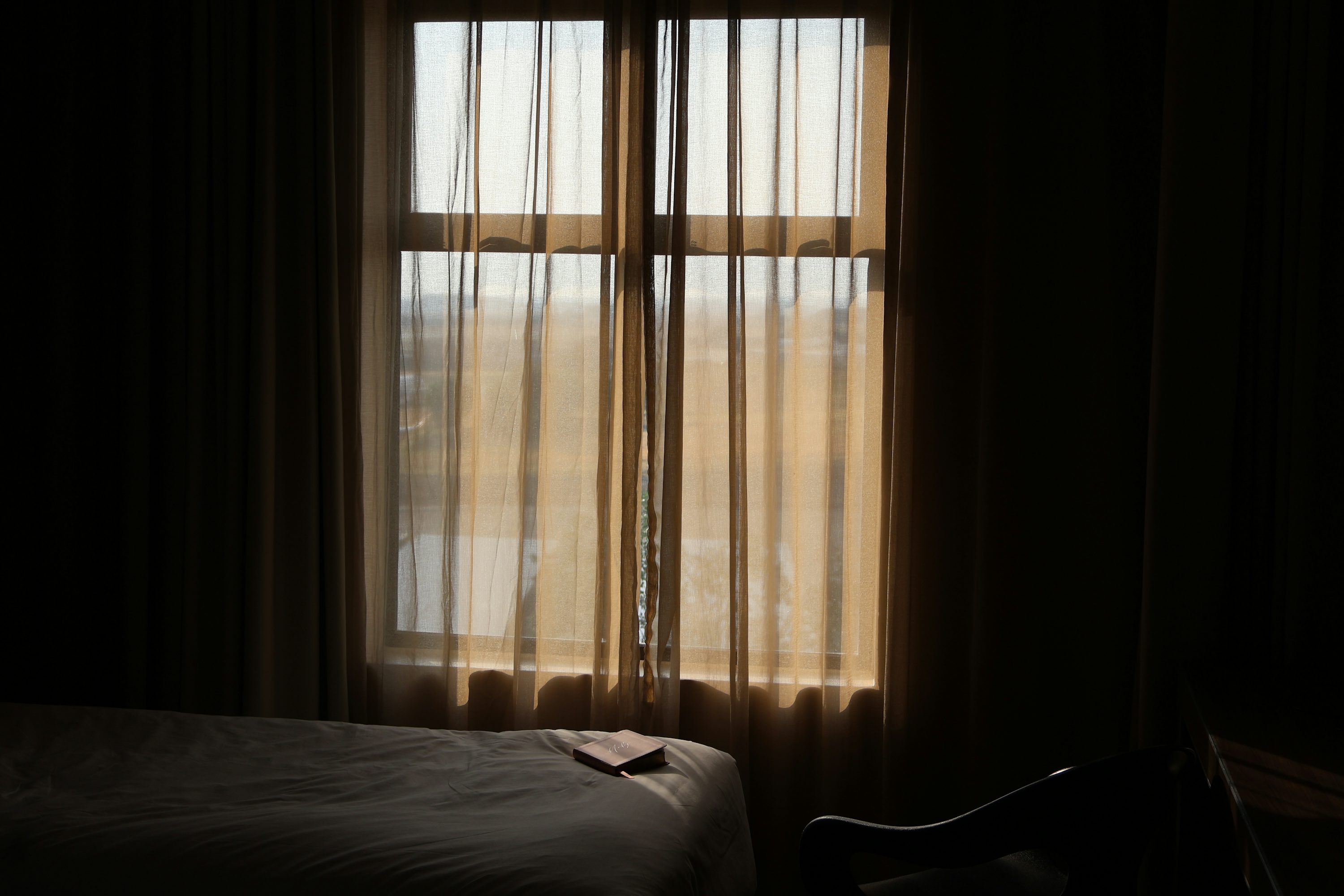 A dim bedroom with soft light through sheer curtains and a book on the bed