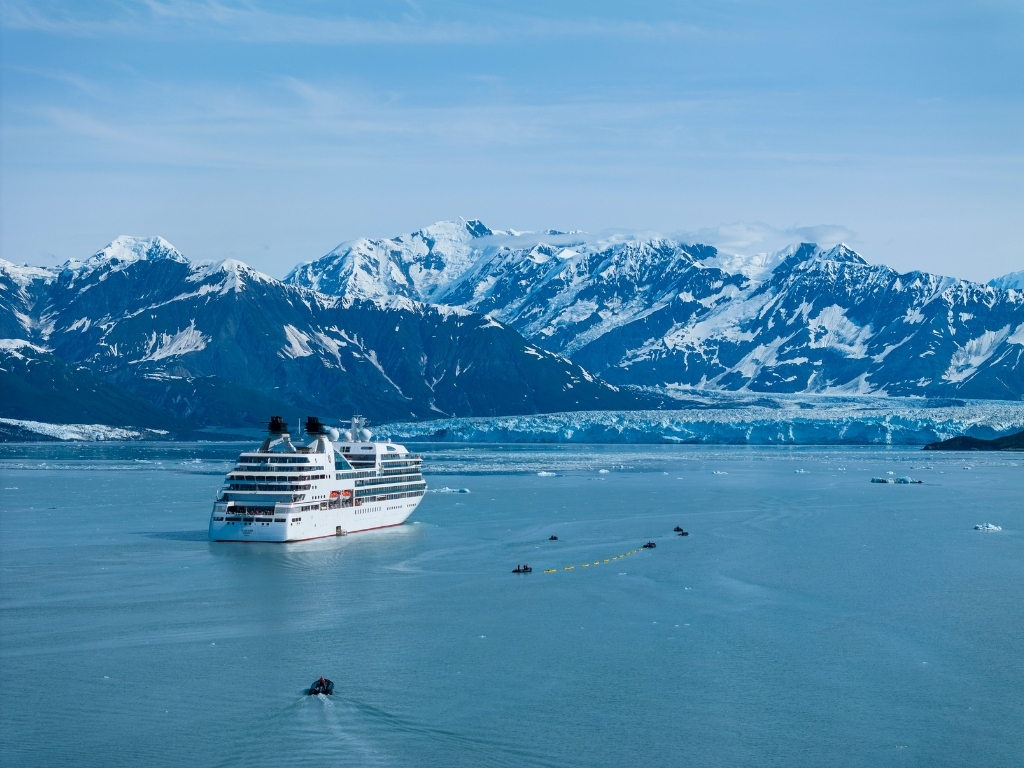 A cruise ship glides across calm Alaskan water in front of snow-capped mountains and a tidewater glacier.